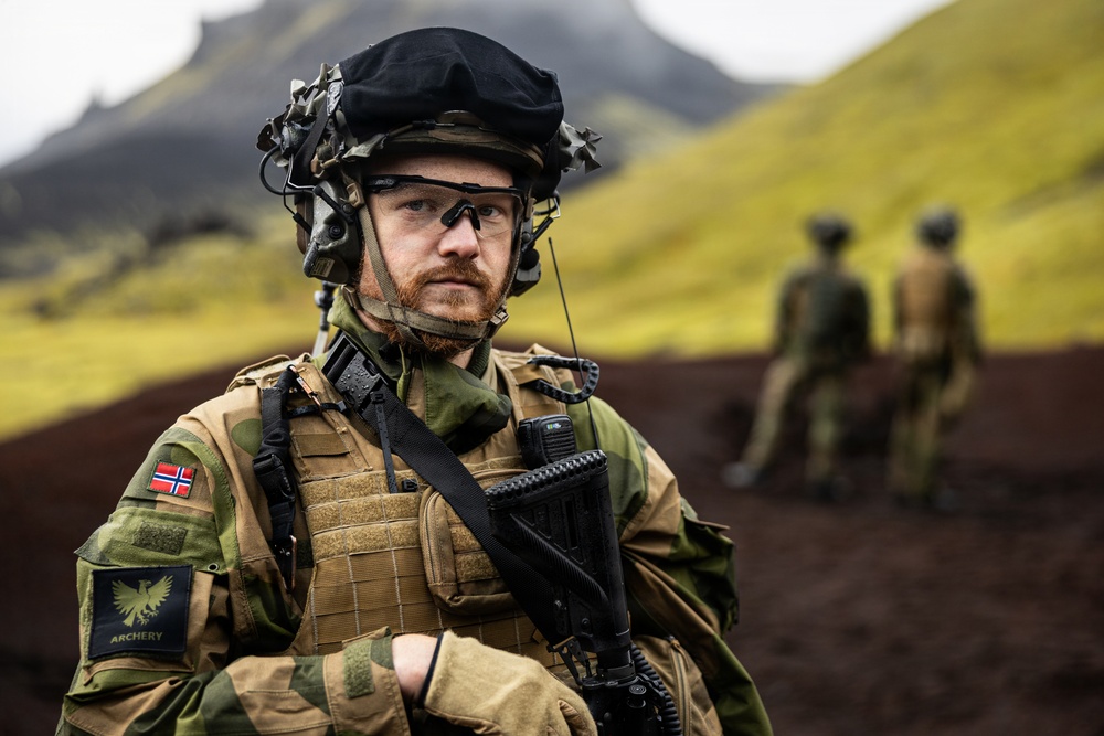 A Norwegian military soldier in full combat gear stands in the foreground of a rugged, volcanic landscape with moss-covered hills in the background. He wears a Norwegian flag patch and an 'ARCHERY' unit patch, equipped with tactical vest, helmet with night-vision mount, communications headset, and a radio. Two additional soldiers are visible out of focus in the background.