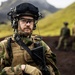 A Norwegian military soldier in full combat gear stands in the foreground of a rugged, volcanic landscape with moss-covered hills in the background. He wears a Norwegian flag patch and an 'ARCHERY' unit patch, equipped with tactical vest, helmet with night-vision mount, communications headset, and a radio. Two additional soldiers are visible out of focus in the background.