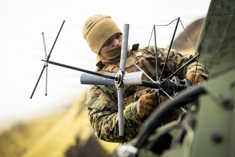 A military service member in camouflage uniform is assembling or adjusting a multi-element antenna system mounted on what appears to be a military vehicle. The individual is wearing cold-weather gear including a tan beanie and face covering, along with tactical gloves. The setting appears to be an outdoor field environment with overcast skies.