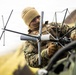 A military service member in camouflage uniform is assembling or adjusting a multi-element antenna system mounted on what appears to be a military vehicle. The individual is wearing cold-weather gear including a tan beanie and face covering, along with tactical gloves. The setting appears to be an outdoor field environment with overcast skies.