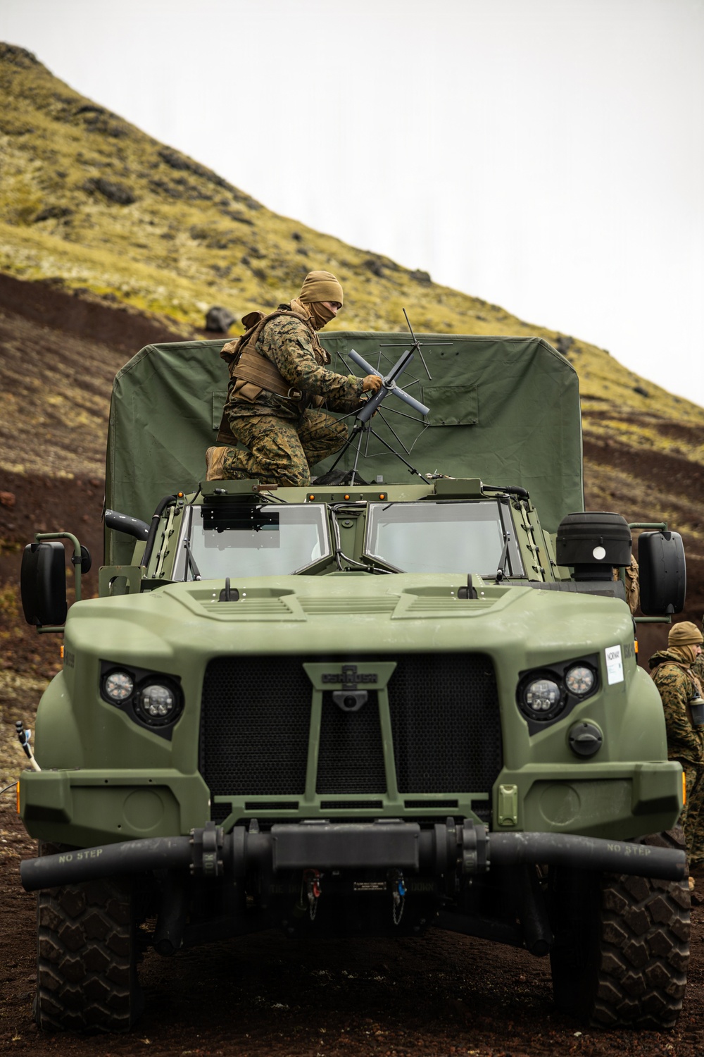 A military service member in MARPAT camouflage and balaclava is positioned on top of an Oshkosh JLTV (Joint Light Tactical Vehicle) painted in olive drab, handling what appears to be a communications or antenna array. The vehicle is parked on rugged terrain with a steep hillside visible in the background. At least one additional service member is visible to the right rear of the vehicle.