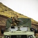 A military service member in MARPAT camouflage and balaclava is positioned on top of an Oshkosh JLTV (Joint Light Tactical Vehicle) painted in olive drab, handling what appears to be a communications or antenna array. The vehicle is parked on rugged terrain with a steep hillside visible in the background. At least one additional service member is visible to the right rear of the vehicle.