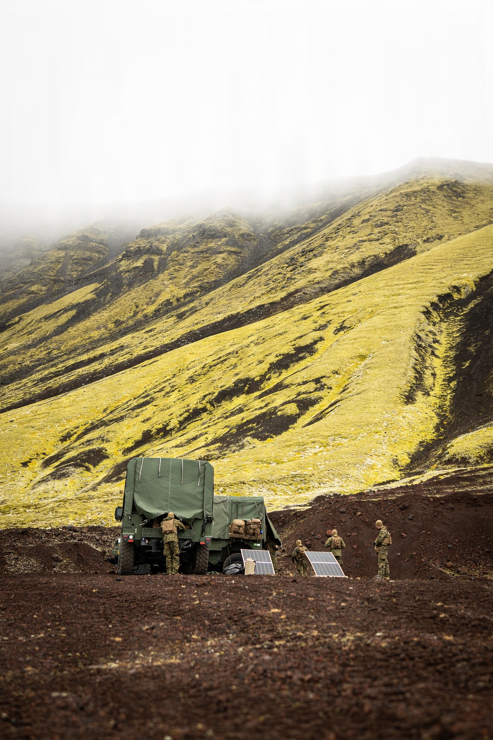 Military personnel are operating in a remote volcanic highland terrain, likely in Iceland based on the distinctive moss-covered lava fields and characteristic landscape. A green canvas-covered military truck is positioned on dark volcanic soil, with several soldiers working around it alongside deployed solar panels. The scene suggests a field operations setup or forward operating base being established in an austere environment.