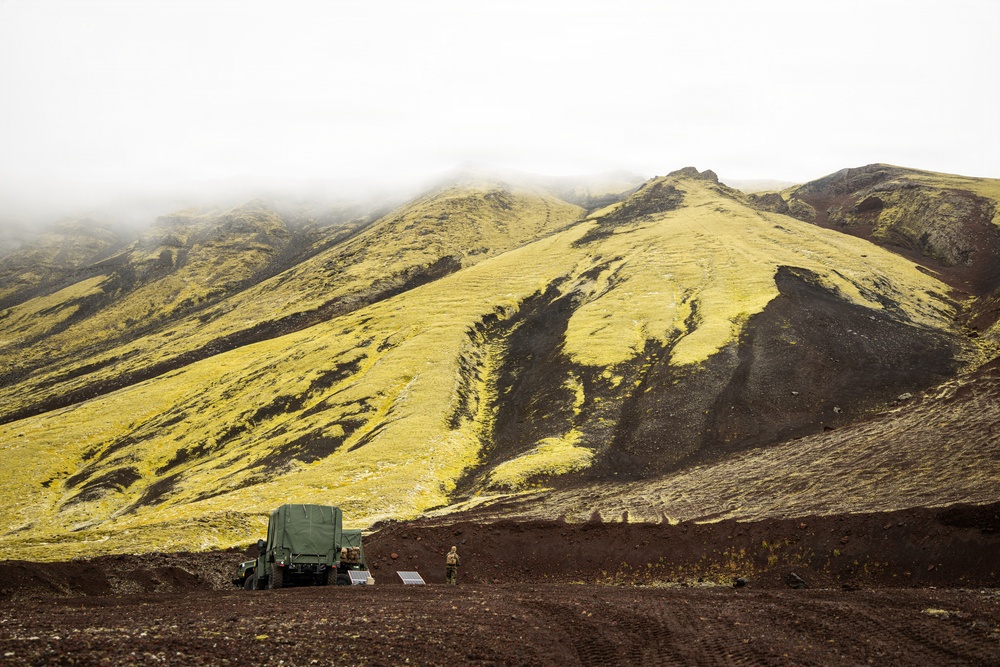 A military-style green cargo truck is parked on dark volcanic terrain at the base of a steep, moss-covered mountain ridge. The landscape features striking yellow-green moss contrasting with black volcanic rock, consistent with Icelandic highland terrain. At least one person is visible standing near the vehicle, and low-lying fog obscures the upper portion of the mountain.