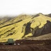 A military-style green cargo truck is parked on dark volcanic terrain at the base of a steep, moss-covered mountain ridge. The landscape features striking yellow-green moss contrasting with black volcanic rock, consistent with Icelandic highland terrain. At least one person is visible standing near the vehicle, and low-lying fog obscures the upper portion of the mountain.