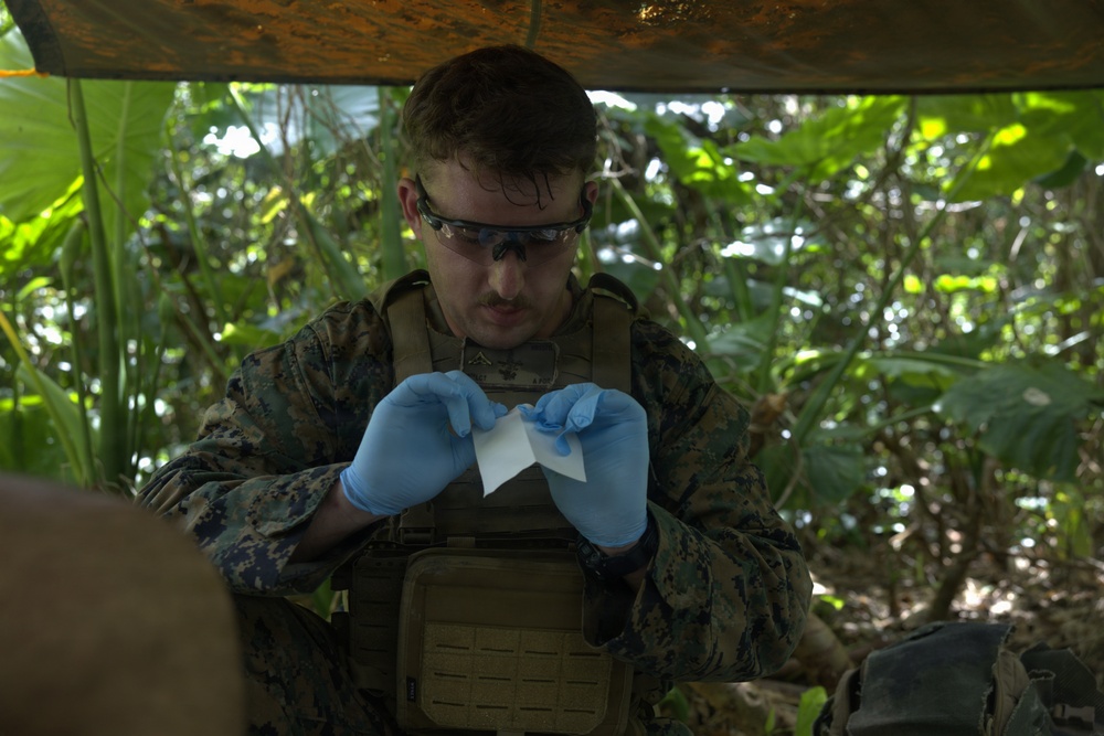 A military service member wearing MARPAT camouflage uniform and tactical vest is handling what appears to be a medical dressing or gauze while wearing blue nitrile gloves. The individual is working under a tarp or canopy in a jungle or tropical environment. The scene suggests a field medical treatment or training exercise.
