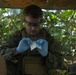 A military service member wearing MARPAT camouflage uniform and tactical vest is handling what appears to be a medical dressing or gauze while wearing blue nitrile gloves. The individual is working under a tarp or canopy in a jungle or tropical environment. The scene suggests a field medical treatment or training exercise.