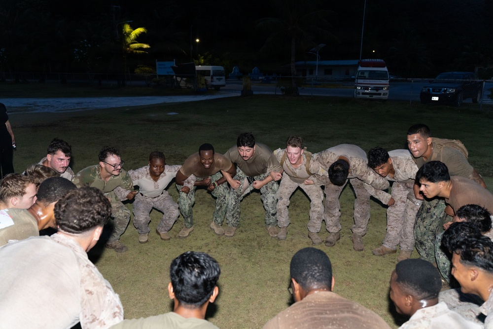 A group of military personnel in camouflage uniforms are gathered in a circle on a grass field at night, appearing to engage in a team-building or physical training exercise. The individuals are leaning inward toward the center of the circle in a coordinated formation. The setting appears to be an outdoor military installation or base, with vehicles and palm trees visible in the background.
