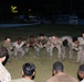 A group of military personnel in camouflage uniforms are gathered in a circle on a grass field at night, appearing to engage in a team-building or physical training exercise. The individuals are leaning inward toward the center of the circle in a coordinated formation. The setting appears to be an outdoor military installation or base, with vehicles and palm trees visible in the background.