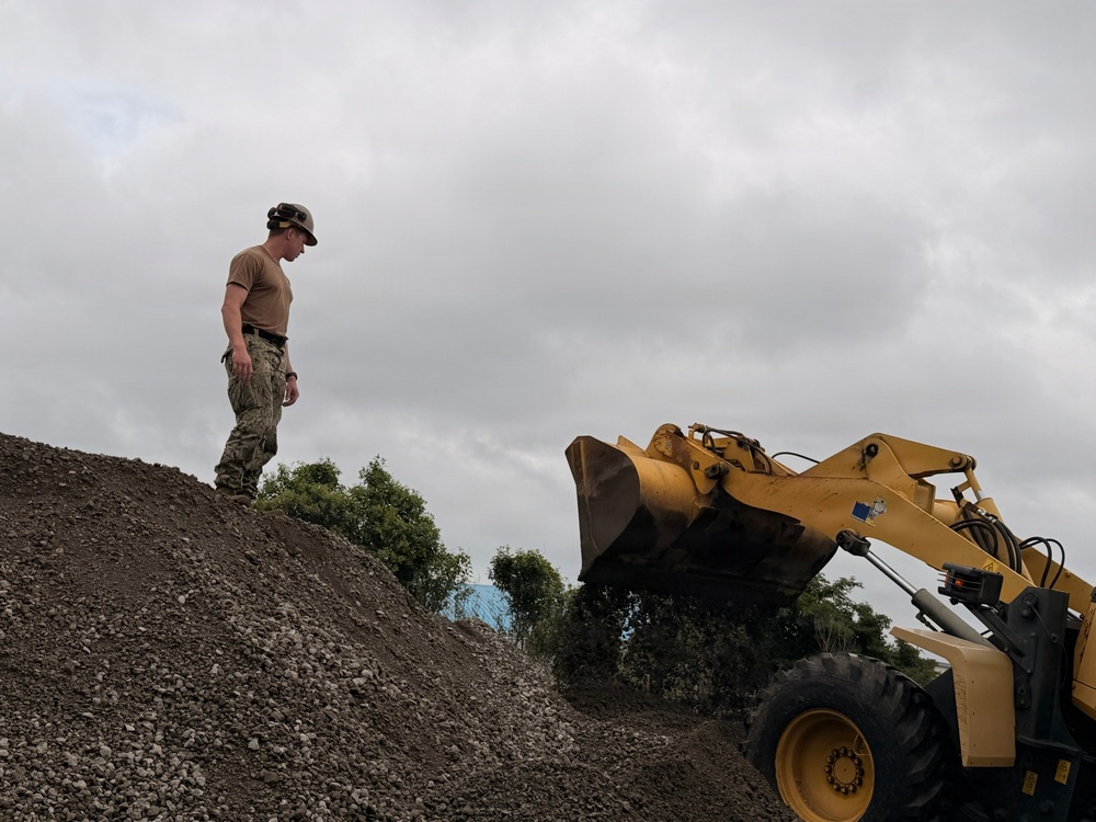 A person in military-style camouflage uniform and hard hat stands atop a large mound of gravel or crushed rock, appearing to supervise or direct operations. A yellow front-end loader (wheel loader) is positioned to the right with its bucket raised, likely engaged in moving or depositing material. The scene appears to be a construction or earthmoving worksite under overcast skies.