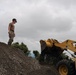 A person in military-style camouflage uniform and hard hat stands atop a large mound of gravel or crushed rock, appearing to supervise or direct operations. A yellow front-end loader (wheel loader) is positioned to the right with its bucket raised, likely engaged in moving or depositing material. The scene appears to be a construction or earthmoving worksite under overcast skies.