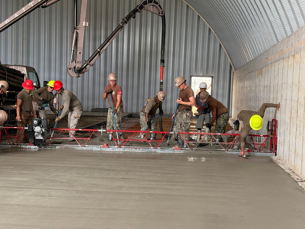A group of workers, appearing to be U.S. Navy Seabees based on their camouflage uniforms, are pouring and leveling a concrete floor inside a large metal arch-roofed building. They are using a mechanical screed (red truss screed) to level the freshly poured concrete, with a concrete pump boom delivering material from outside. The work is actively in progress with multiple personnel managing the pour and finishing process.