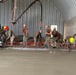 A group of workers, appearing to be U.S. Navy Seabees based on their camouflage uniforms, are pouring and leveling a concrete floor inside a large metal arch-roofed building. They are using a mechanical screed (red truss screed) to level the freshly poured concrete, with a concrete pump boom delivering material from outside. The work is actively in progress with multiple personnel managing the pour and finishing process.