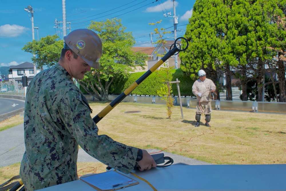 Two personnel, one in military camouflage uniform and one in a tan work suit, are conducting what appears to be an explosive ordnance disposal (EOD) or utility detection exercise on a military installation. The foreground figure wearing a hard hat with a unit patch is operating equipment at a table, while the background figure holds a detection wand or probe connected by a cable. The setting appears to be a well-maintained outdoor area consistent with a U.S. military base in Japan.