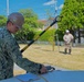 Two personnel, one in military camouflage uniform and one in a tan work suit, are conducting what appears to be an explosive ordnance disposal (EOD) or utility detection exercise on a military installation. The foreground figure wearing a hard hat with a unit patch is operating equipment at a table, while the background figure holds a detection wand or probe connected by a cable. The setting appears to be a well-maintained outdoor area consistent with a U.S. military base in Japan.