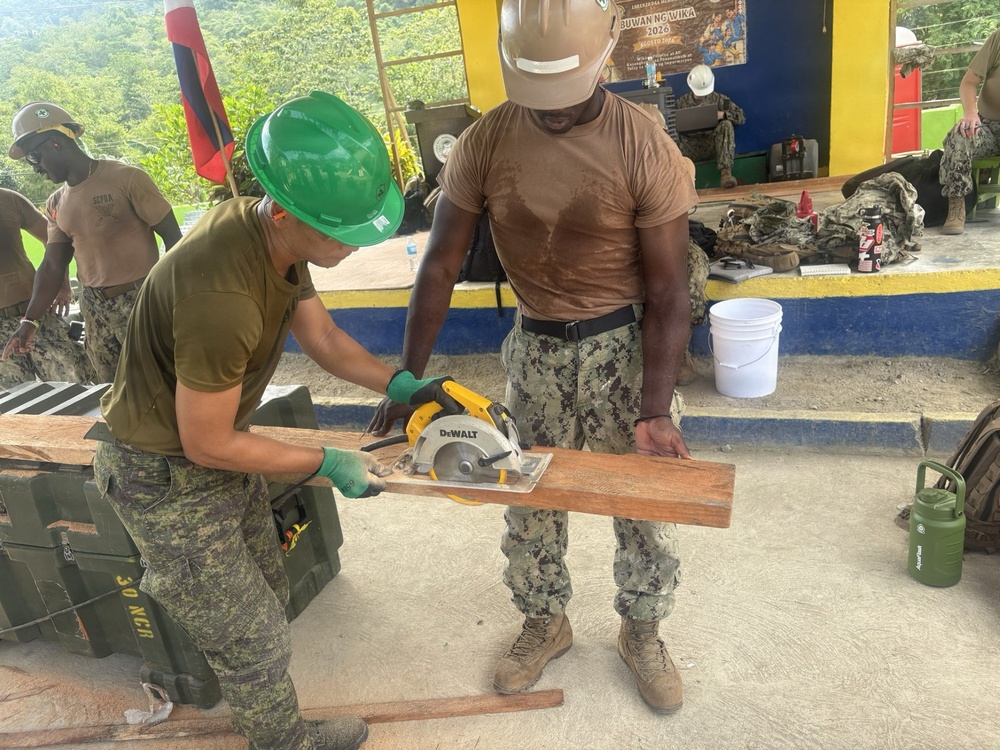 Two military personnel are engaged in construction work, with one soldier wearing a green hard hat using a DeWalt circular saw to cut a wooden plank while the other steadies the board. The setting appears to be an outdoor covered area, likely in the Philippines based on the 'Buwan ng Wika 2026' banner visible in the background. The personnel appear to represent a joint military operation, with one wearing Philippine Army camouflage and the other in US Navy digital camouflage (NWU).