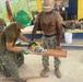 Two military personnel are engaged in construction work, with one soldier wearing a green hard hat using a DeWalt circular saw to cut a wooden plank while the other steadies the board. The setting appears to be an outdoor covered area, likely in the Philippines based on the 'Buwan ng Wika 2026' banner visible in the background. The personnel appear to represent a joint military operation, with one wearing Philippine Army camouflage and the other in US Navy digital camouflage (NWU).