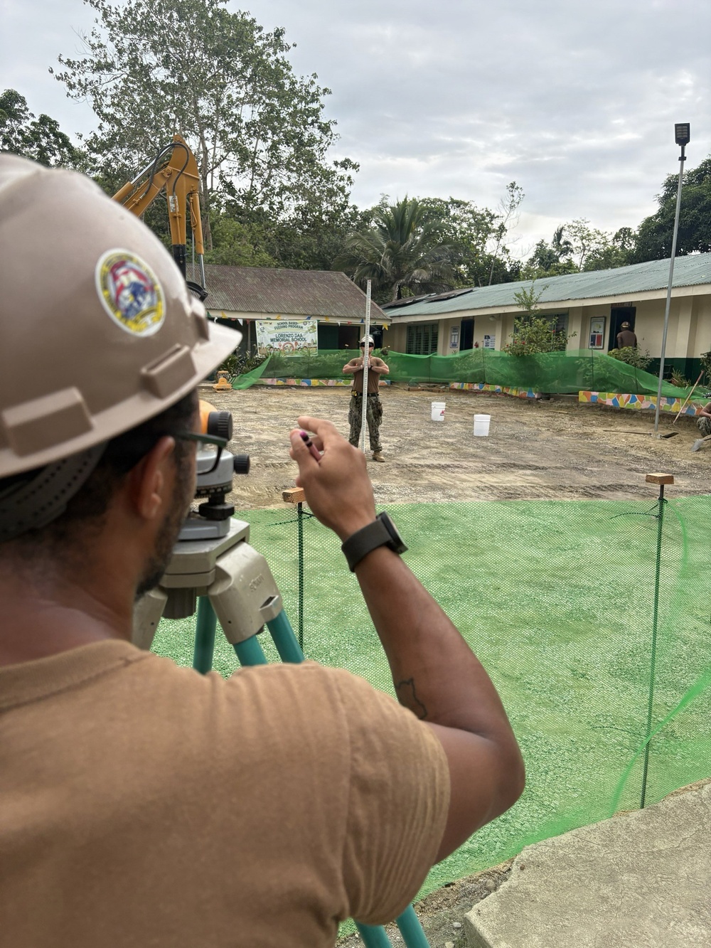 A construction worker wearing a tan hard hat with an emblem is operating a surveying instrument (optical level/theodolite) on a tripod at an active construction site. In the background, a second worker in military-style clothing holds a surveying rod/staff while standing in a cleared dirt area. The site appears to be adjacent to a school building, visible in the background with a sign referencing 'Lorenzo Daa Memorial School' and a 'School-Based Feeding Program' banner.