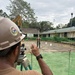 A construction worker wearing a tan hard hat with an emblem is operating a surveying instrument (optical level/theodolite) on a tripod at an active construction site. In the background, a second worker in military-style clothing holds a surveying rod/staff while standing in a cleared dirt area. The site appears to be adjacent to a school building, visible in the background with a sign referencing 'Lorenzo Daa Memorial School' and a 'School-Based Feeding Program' banner.