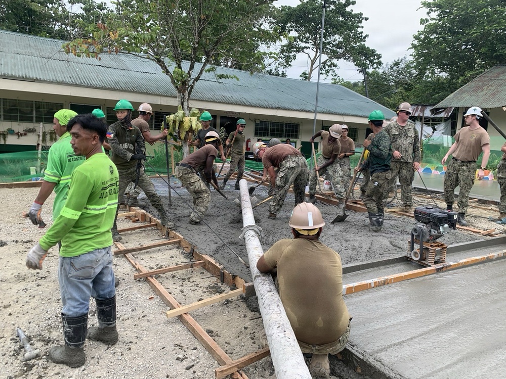 A group of military personnel and civilian workers are engaged in a concrete pouring and leveling operation, likely at a school or community facility. Workers in camouflage uniforms, green hard hats, and civilian clothing are collaborating to spread and screed fresh concrete within wooden formwork. The setting appears to be in a tropical region, possibly the Philippines, based on vegetation, architecture, and uniform markings.