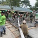 A group of military personnel and civilian workers are engaged in a concrete pouring and leveling operation, likely at a school or community facility. Workers in camouflage uniforms, green hard hats, and civilian clothing are collaborating to spread and screed fresh concrete within wooden formwork. The setting appears to be in a tropical region, possibly the Philippines, based on vegetation, architecture, and uniform markings.