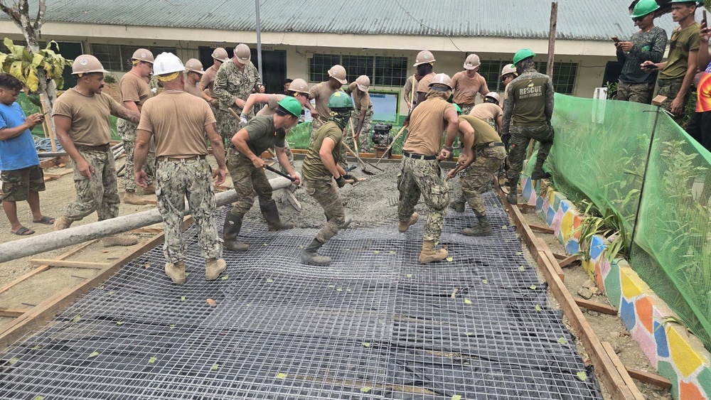 A large group of military personnel in uniform are engaged in a concrete pouring and spreading operation, likely as part of a humanitarian construction project. Workers wearing various colored hard hats (tan, white, and green) are using shovels and rakes to spread wet concrete over a reinforced steel mesh grid. The setting appears to be near a school or community building, suggesting a civic action or engineering assistance mission.