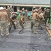 A large group of military personnel in uniform are engaged in a concrete pouring and spreading operation, likely as part of a humanitarian construction project. Workers wearing various colored hard hats (tan, white, and green) are using shovels and rakes to spread wet concrete over a reinforced steel mesh grid. The setting appears to be near a school or community building, suggesting a civic action or engineering assistance mission.