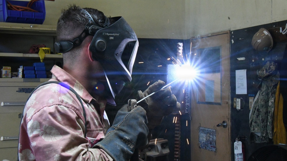 A welder is actively performing a welding operation in what appears to be a workshop or maintenance facility. The person is wearing a 3M Speedglas auto-darkening welding helmet and heavy-duty leather welding gloves, with bright welding sparks and arc light visible. The background shows shelving units, storage cabinets, and what appears to be military-style gear hanging on a door.