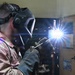 A welder is actively performing a welding operation in what appears to be a workshop or maintenance facility. The person is wearing a 3M Speedglas auto-darkening welding helmet and heavy-duty leather welding gloves, with bright welding sparks and arc light visible. The background shows shelving units, storage cabinets, and what appears to be military-style gear hanging on a door.