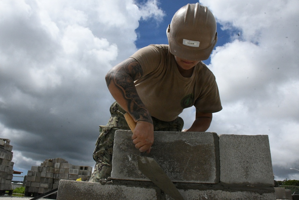 A worker wearing a tan hard hat labeled 'HORN' and camouflage pants is laying concrete masonry blocks (CMUs) outdoors. The individual has a full sleeve tattoo on their right arm and is using a masonry trowel to apply mortar between blocks. The low-angle shot frames the worker against a dramatic partly cloudy sky.
