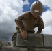 A worker wearing a tan hard hat labeled 'HORN' and camouflage pants is laying concrete masonry blocks (CMUs) outdoors. The individual has a full sleeve tattoo on their right arm and is using a masonry trowel to apply mortar between blocks. The low-angle shot frames the worker against a dramatic partly cloudy sky.