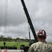 A military service member wearing US Navy digital camouflage (NWU Type I) and a red hard hat with hearing protection is observing a crane operation at what appears to be a military installation. A large olive-drab military crane is extended at an angle over a paved area, with additional military vehicles visible in the background. The setting appears to be a tropical or subtropical location based on the lush green vegetation visible beyond the perimeter fence.