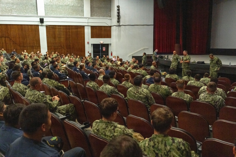 A large group of military personnel, predominantly wearing U.S. Navy digital camouflage (NWU Type III) uniforms, are seated in an auditorium or theater-style venue. Several uniformed individuals are standing near the stage area, appearing to address or brief the assembled group. The setting suggests a formal military all-hands meeting, briefing, or command presentation.