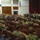 A large group of military personnel, predominantly wearing U.S. Navy digital camouflage (NWU Type III) uniforms, are seated in an auditorium or theater-style venue. Several uniformed individuals are standing near the stage area, appearing to address or brief the assembled group. The setting suggests a formal military all-hands meeting, briefing, or command presentation.