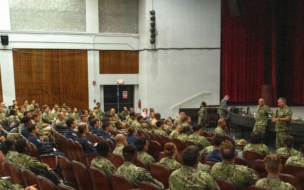 A large group of military personnel, predominantly wearing U.S. Navy digital camouflage (NWU Type III) uniforms, are seated in an auditorium or theater-style venue. Several uniformed individuals are standing near the stage area, appearing to address or brief the assembled group. The setting suggests a formal military all-hands meeting, briefing, or command presentation.