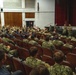 A large group of military personnel, predominantly wearing U.S. Navy digital camouflage (NWU Type III) uniforms, are seated in an auditorium or theater-style venue. Several uniformed individuals are standing near the stage area, appearing to address or brief the assembled group. The setting suggests a formal military all-hands meeting, briefing, or command presentation.