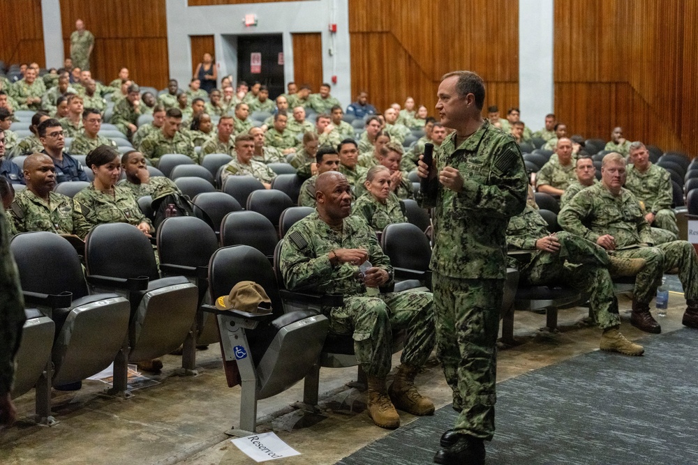 A senior military officer in U.S. Navy Working Uniform (NWU Type III camouflage) addresses a large audience of uniformed naval personnel in an auditorium or theater setting. The speaker is holding a microphone and appears to be conducting an all-hands call or briefing. The audience consists of dozens of sailors seated in rows of gray auditorium chairs.