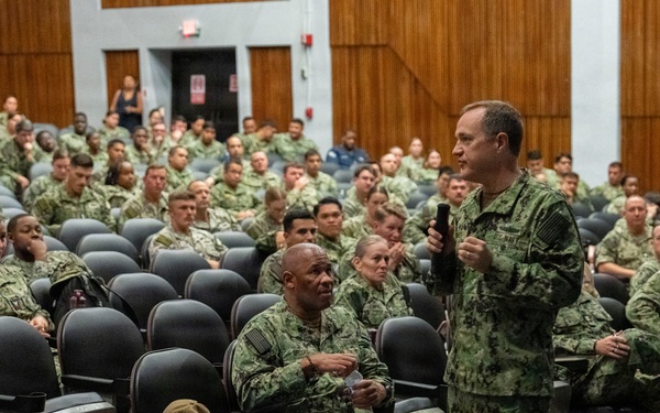 A senior military officer in U.S. Navy Working Uniform (NWU Type III camouflage) addresses a large audience of uniformed naval personnel in an auditorium or theater setting. The speaker is holding a microphone and appears to be conducting an all-hands call or briefing. The audience consists of dozens of sailors seated in rows of gray auditorium chairs.