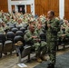 A senior military officer in U.S. Navy Working Uniform (NWU Type III camouflage) addresses a large audience of uniformed naval personnel in an auditorium or theater setting. The speaker is holding a microphone and appears to be conducting an all-hands call or briefing. The audience consists of dozens of sailors seated in rows of gray auditorium chairs.