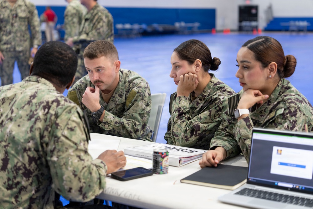 Four U.S. Navy personnel in NWU Type III camouflage uniforms are seated around a white table in what appears to be a gymnasium or large indoor facility. Three sailors face a fourth sailor whose back is to the camera, suggesting a briefing, counseling, or administrative session. A laptop, tablet, books, and documents are visible on the table, indicating a formal or educational meeting.