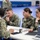 Four U.S. Navy personnel in NWU Type III camouflage uniforms are seated around a white table in what appears to be a gymnasium or large indoor facility. Three sailors face a fourth sailor whose back is to the camera, suggesting a briefing, counseling, or administrative session. A laptop, tablet, books, and documents are visible on the table, indicating a formal or educational meeting.