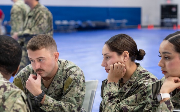 Four U.S. Navy personnel in NWU Type III camouflage uniforms are seated around a white table in what appears to be a gymnasium or large indoor facility. Three sailors face a fourth sailor whose back is to the camera, suggesting a briefing, counseling, or administrative session. A laptop, tablet, books, and documents are visible on the table, indicating a formal or educational meeting.