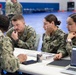 Four U.S. Navy personnel in NWU Type III camouflage uniforms are seated around a white table in what appears to be a gymnasium or large indoor facility. Three sailors face a fourth sailor whose back is to the camera, suggesting a briefing, counseling, or administrative session. A laptop, tablet, books, and documents are visible on the table, indicating a formal or educational meeting.