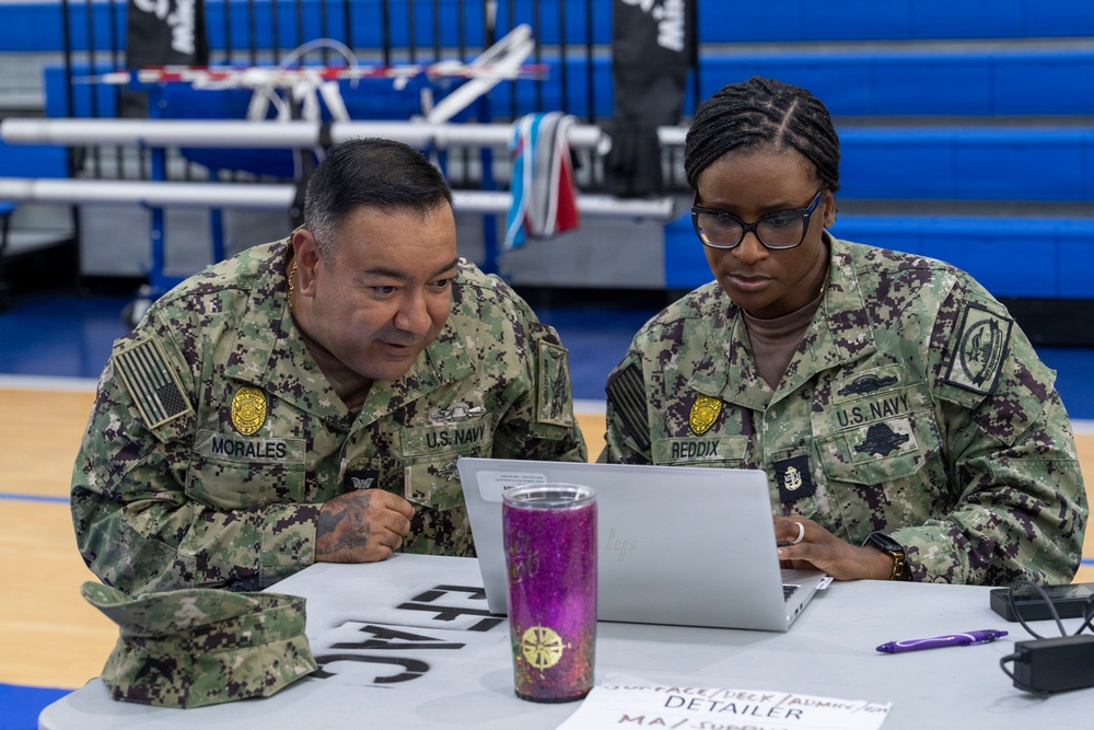 Two U.S. Navy personnel in NWU Type III camouflage uniforms are seated at a table reviewing information on a laptop computer. The name tapes visible identify them as 'MORALES' (left) and 'REDDIX' (right), both wearing U.S. Navy insignia and rank devices. They appear to be working at an administrative or detailer station inside what appears to be a gymnasium or sports facility.