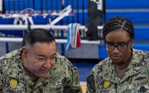 Two U.S. Navy personnel in NWU Type III camouflage uniforms are seated at a table reviewing information on a laptop computer. The name tapes visible identify them as 'MORALES' (left) and 'REDDIX' (right), both wearing U.S. Navy insignia and rank devices. They appear to be working at an administrative or detailer station inside what appears to be a gymnasium or sports facility.