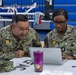 Two U.S. Navy personnel in NWU Type III camouflage uniforms are seated at a table reviewing information on a laptop computer. The name tapes visible identify them as 'MORALES' (left) and 'REDDIX' (right), both wearing U.S. Navy insignia and rank devices. They appear to be working at an administrative or detailer station inside what appears to be a gymnasium or sports facility.