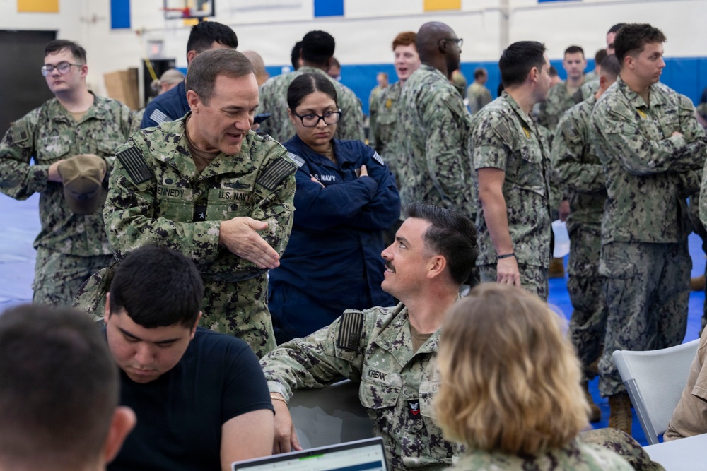 A group of U.S. Navy personnel are gathered in what appears to be a gymnasium or large indoor facility, likely during a briefing, processing event, or administrative muster. A senior officer with the name tape 'KENNEDY' and a star insignia is engaged in conversation with another officer named 'KRENK' who is seated at a table with a laptop. The setting suggests an organized military event with multiple sailors in Navy Working Uniform (NWU Type III) camouflage pattern.