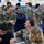 A group of U.S. Navy personnel are gathered in what appears to be a gymnasium or large indoor facility, likely during a briefing, processing event, or administrative muster. A senior officer with the name tape 'KENNEDY' and a star insignia is engaged in conversation with another officer named 'KRENK' who is seated at a table with a laptop. The setting suggests an organized military event with multiple sailors in Navy Working Uniform (NWU Type III) camouflage pattern.