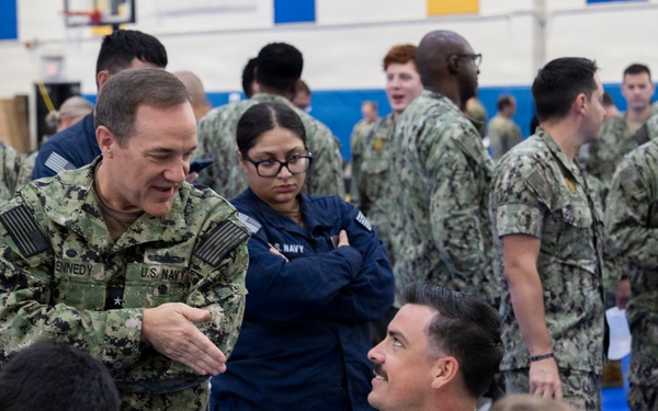 A group of U.S. Navy personnel are gathered in what appears to be a gymnasium or large indoor facility, likely during a briefing, processing event, or administrative muster. A senior officer with the name tape 'KENNEDY' and a star insignia is engaged in conversation with another officer named 'KRENK' who is seated at a table with a laptop. The setting suggests an organized military event with multiple sailors in Navy Working Uniform (NWU Type III) camouflage pattern.