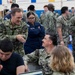 A group of U.S. Navy personnel are gathered in what appears to be a gymnasium or large indoor facility, likely during a briefing, processing event, or administrative muster. A senior officer with the name tape 'KENNEDY' and a star insignia is engaged in conversation with another officer named 'KRENK' who is seated at a table with a laptop. The setting suggests an organized military event with multiple sailors in Navy Working Uniform (NWU Type III) camouflage pattern.